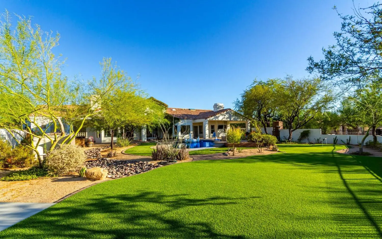 A spacious backyard in Peoria AZ features lush artificial grass, desert landscaping, trees, and a large house in the background. Enjoy a covered patio and a pool surrounded by plants under a clear blue sky.