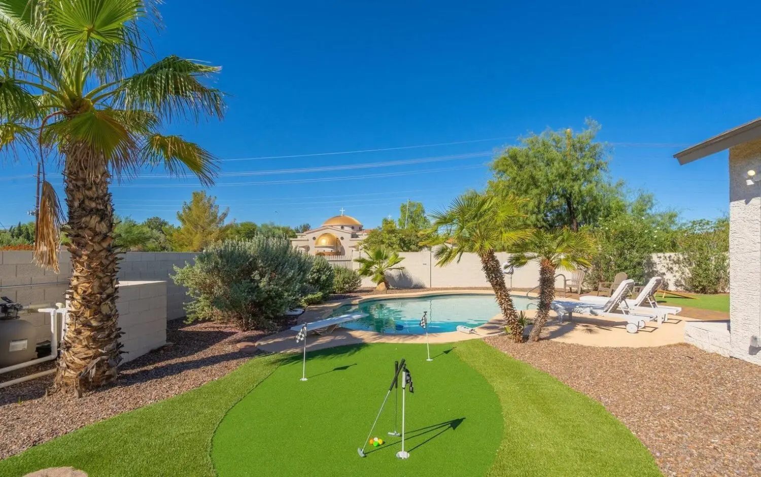 A backyard with a small artificial putting green, golf flags, palm trees, lounge chairs, and a swimming pool under a clear blue sky features premium synthetic turf Glendale for lasting beauty.