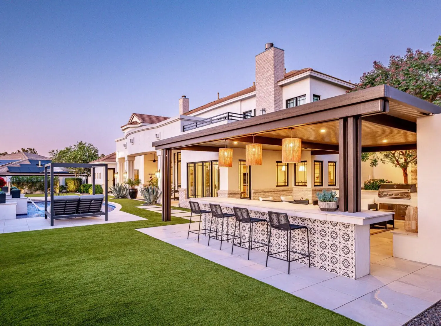 Modern backyard in Litchfield Park AZ with a covered outdoor kitchen and bar, patterned tile accents, hanging lights, and bar stools. Features include a pool, lush turf installation, and a large white two-story house in the sunset glow.
