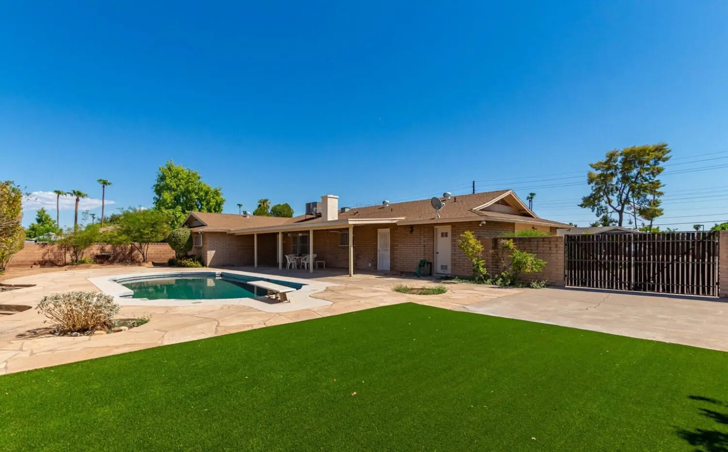 Backyard with synthetic turf, stone patio, small pool, and a single-story brick house featuring a covered patio. A few trees and shrubs line the wooden fence, all under a clear blue sky—perfect showcase for expert artificial grass installation.
