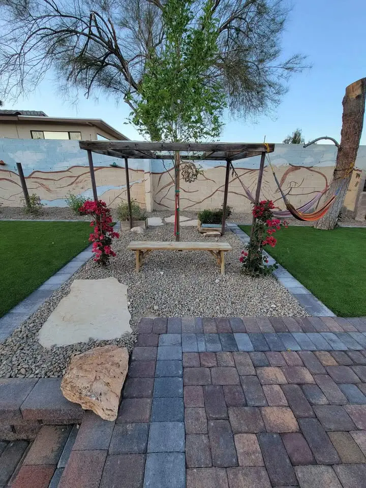 A landscaped backyard with a stone path leading to a bench under a pergola decorated with red flowering vines. Lush synthetic turf Glendale enhances the green grass, while a hammock rests on the right and a mural decorates the low wall in back.