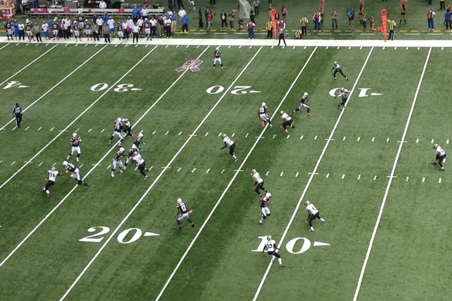 Aerial view of an American football game on synthetic turf, with a quarterback preparing to throw as offensive and defensive players are spread out near the 20- and 10-yard lines.